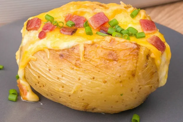 Batata assada recheada com queijo derretido, pedaços de bacon crocante e cebolinha, servida em um prato cinza.