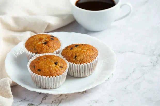 Três bolinhos de fubá servidos em forminhas brancas sobre um prato de porcelana, acompanhados por uma xícara de café em fundo claro.