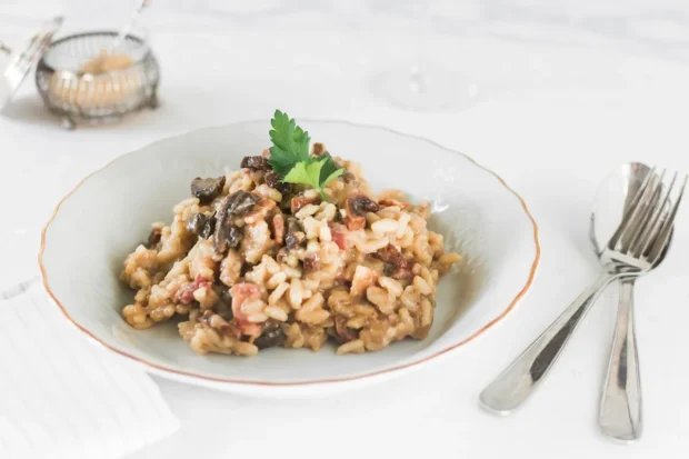 Prato de risotto de funghi e bacon servido em uma tigela branca, decorado com uma folha de salsa, acompanhado por talheres de inox sobre uma mesa clara.