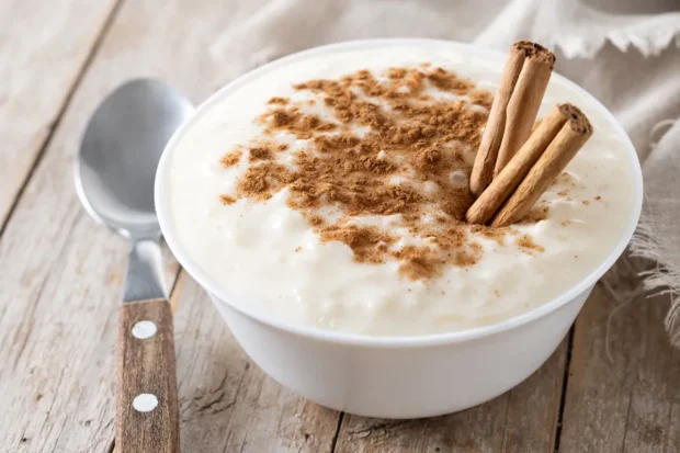  Tigela de arroz doce cremoso polvilhado com canela em pó e decorado com paus de canela, servida sobre uma mesa rústica.