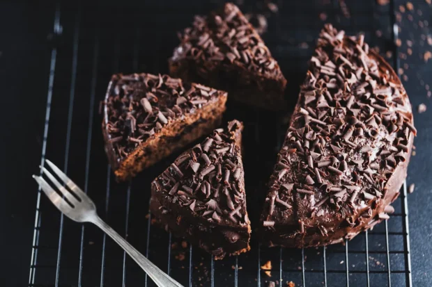 Um bolo de chocolate decorado com raspas de chocolate sobre uma grade de resfriamento, com uma fatia cortada e um garfo ao lado.