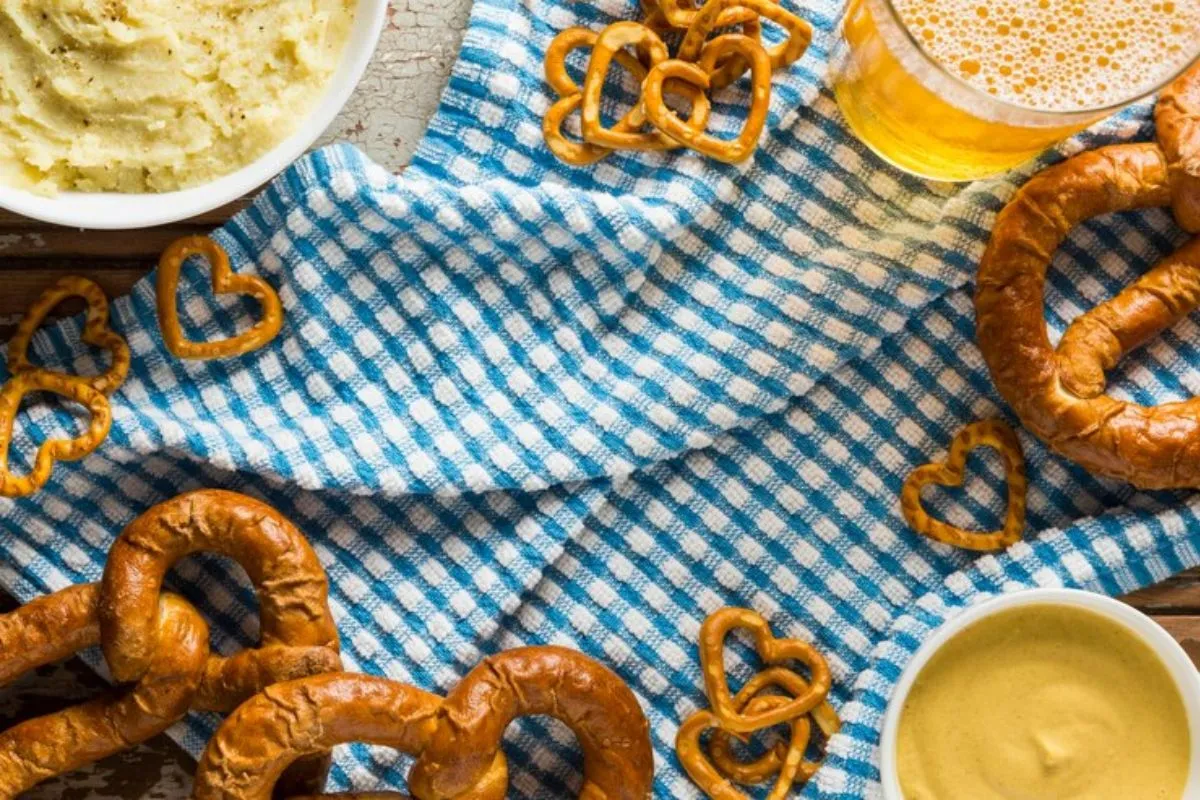 Pretzels e mini pretzels com mostarda e cerveja sobre uma toalha xadrez azul e branca.