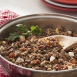 Carne moída refogada com cebola, tomate e ovo cozido em uma frigideira de aço inox, com colher de pau.