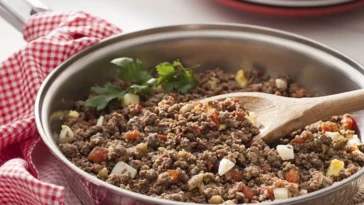 Carne moída refogada com cebola, tomate e ovo cozido em uma frigideira de aço inox, com colher de pau.