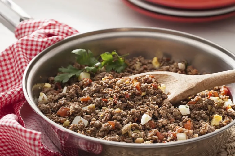 Carne moída refogada com cebola, tomate e ovo cozido em uma frigideira de aço inox, com colher de pau.