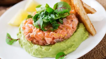 Prato com tartar de salmão fresco sobre creme de abacate, decorado com folhas verdes, acompanhado de torradas e fatias de limão.