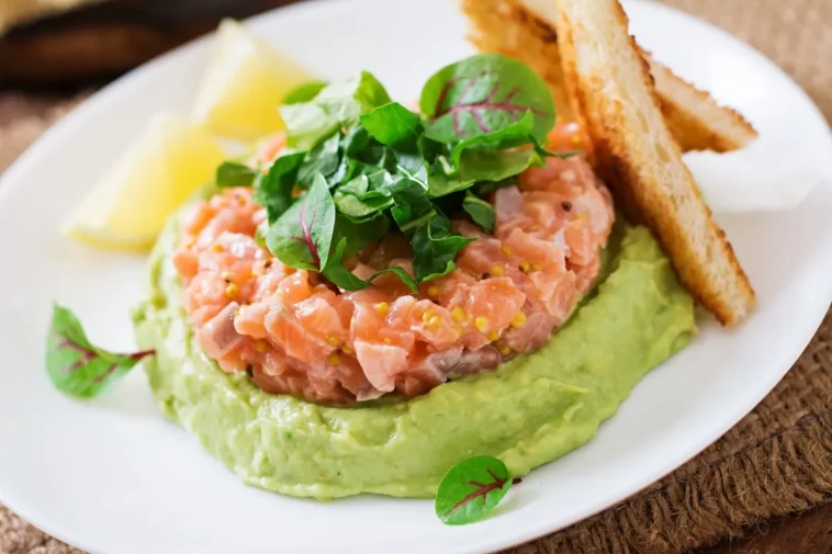 Prato com tartar de salmão fresco sobre creme de abacate, decorado com folhas verdes, acompanhado de torradas e fatias de limão.