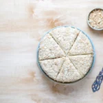 Bolo de tapioca granulado servido em pedaços triangulares, acompanhado de uma tigela com granulado e uma espátula decorada sobre uma mesa de madeira clara.