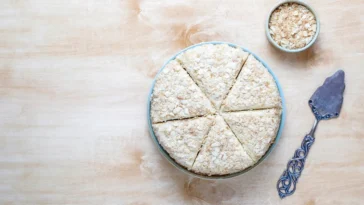 Bolo de tapioca granulado servido em pedaços triangulares, acompanhado de uma tigela com granulado e uma espátula decorada sobre uma mesa de madeira clara.