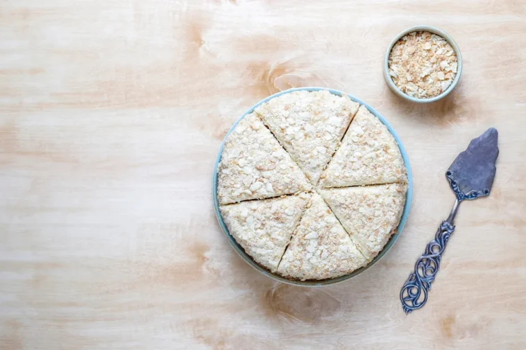 Bolo de tapioca granulado servido em pedaços triangulares, acompanhado de uma tigela com granulado e uma espátula decorada sobre uma mesa de madeira clara.