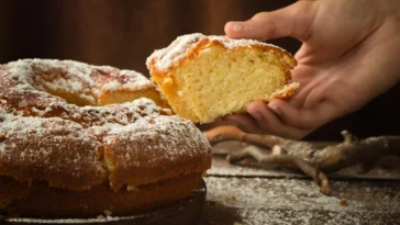 Bolo caseiro fofinho sem fermento coberto com açúcar de confeiteiro, com uma mão segurando uma fatia recém-cortada.