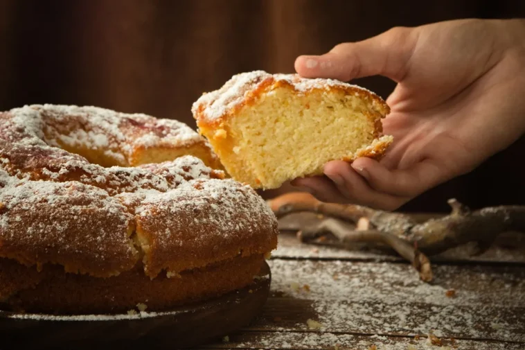 Bolo caseiro fofinho sem fermento coberto com açúcar de confeiteiro, com uma mão segurando uma fatia recém-cortada.