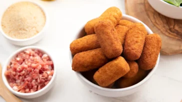 Tigela cheia de croquetes de carne dourados e crocantes, cercada por ingredientes como bacon picado, farinha de rosca e folhas frescas.