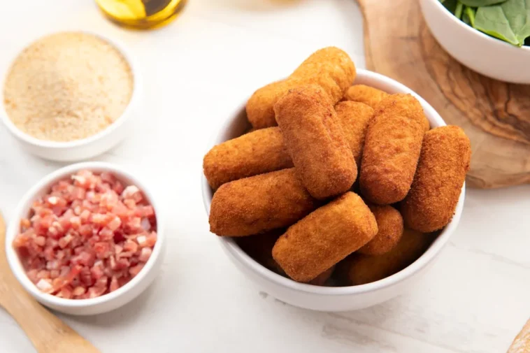 Tigela cheia de croquetes de carne dourados e crocantes, cercada por ingredientes como bacon picado, farinha de rosca e folhas frescas.