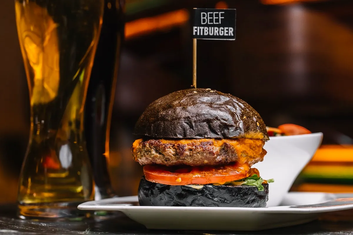 Hambúrguer artesanal com pão preto, queijo derretido, tomate fresco e folhas verdes, servido em um prato branco com uma bandeira indicando "Beef Fitburger".