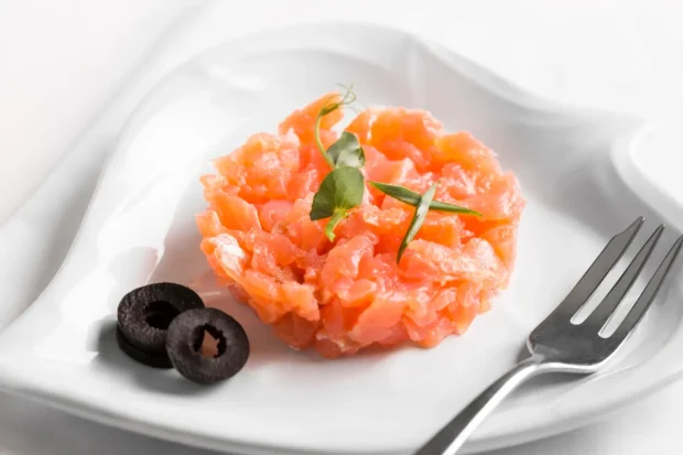 Tartar de salmão fresco em um prato branco em formato de coração, decorado com folhas verdes e acompanhado por fatias de azeitona preta.