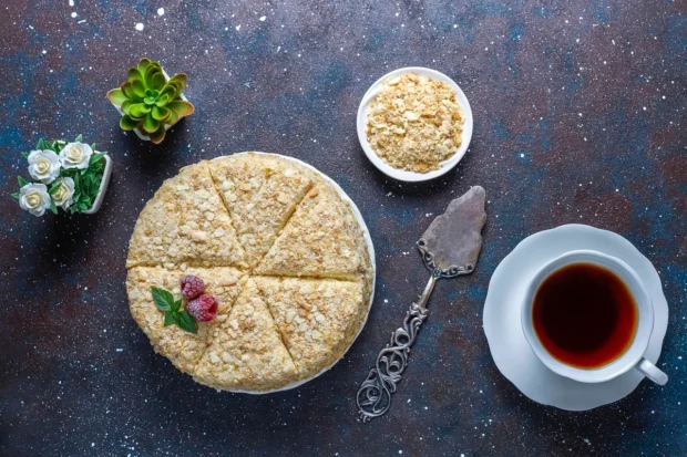 Bolo de tapioca granulado decorado com framboesas e folhas de hortelã, acompanhado por uma xícara de chá, uma tigela com granulado e uma espátula vintage, em uma mesa decorativa.