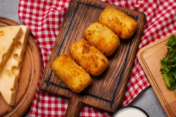 Quatro croquetes dourados dispostos sobre uma tábua de madeira rústica, acompanhados de pedaços de queijo e folhas de alface em um fundo com toalha xadrez vermelha e branca.