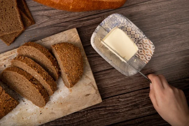 Pão integral com sementes fatiado em uma tábua ao lado de um prato com manteiga caseira sendo cortada, tudo disposto sobre uma mesa de madeira rústica.