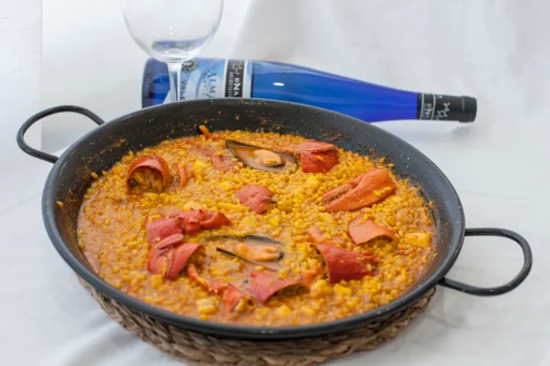 Prato de paella de frutos do mar servido em panela típica, acompanhado por uma garrafa de vinho azul e uma taça.