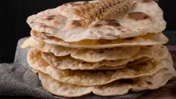 Pilha de pães sírios frescos, levemente tostados, com decoração de espigas de trigo no topo, dispostos sobre um pano rústico.