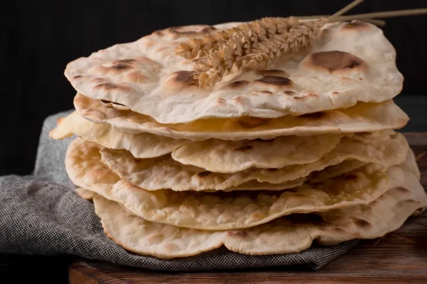 Pilha de pães sírios frescos, levemente tostados, com decoração de espigas de trigo no topo, dispostos sobre um pano rústico.