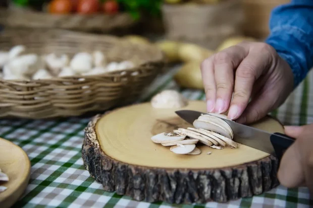 Mãos cortando cogumelos frescos em fatias finas sobre uma tábua rústica de madeira, com cestos de vegetais ao fundo.