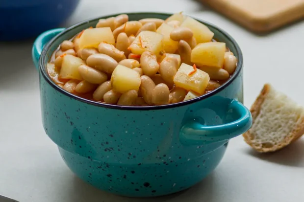 Prato de feijão branco cozido com pedaços de batata e cenoura, servido em uma tigela azul rústica com alça, acompanhado por um pedaço de pão ao fundo.