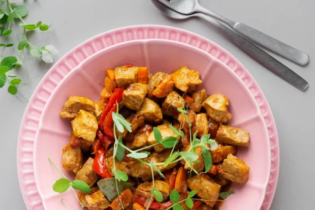 Prato vegano de tofu salteado com legumes, servido em um prato rosa, decorado com brotos verdes frescos e acompanhado de talheres prateados sobre uma mesa cinza.