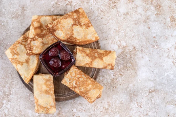 Crepes dobrados em diferentes formatos, dispostos em uma bandeja de madeira, acompanhados de uma tigela com geleia de morango e frutas inteiras.