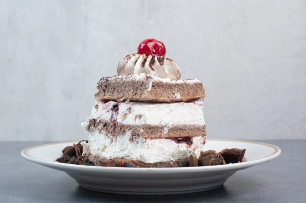 Fatia de torta de chocolate decorada com chantilly e uma cereja no topo, servida em um prato branco com borda dourada.