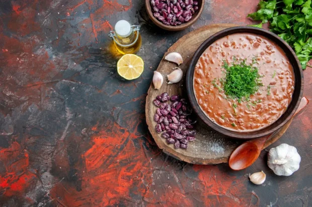 Tigela de sopa de feijão em prato rústico decorado com ervas frescas, cercada por ingredientes como feijões secos, alho, limão e azeite, sobre uma superfície com tons quentes.