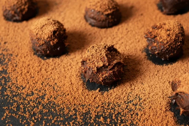 Trufas de chocolate caseiras cobertas com cacau em pó sobre uma superfície preta, com cacau espalhado ao redor.