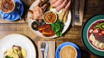 Mesa rústica com pratos variados de brunch, incluindo café com leite, tigela de frutas, prato com ovos e legumes, e uma xícara de cappuccino.