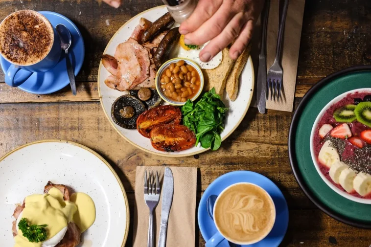 Mesa rústica com pratos variados de brunch, incluindo café com leite, tigela de frutas, prato com ovos e legumes, e uma xícara de cappuccino.