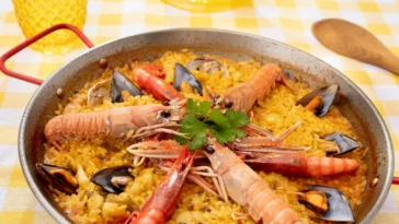 Paella de frutos do mar servida em uma panela típica com camarões, mexilhões e ervas frescas, sobre uma toalha amarela e branca.