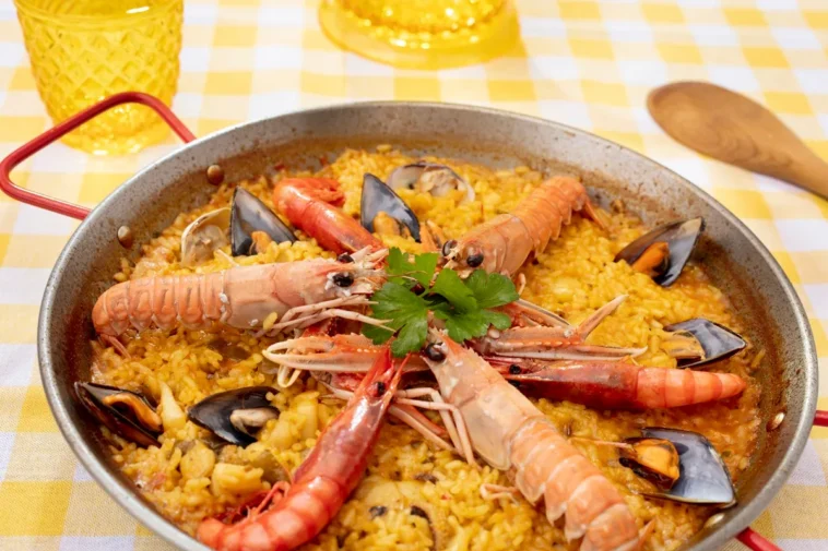 Paella de frutos do mar servida em uma panela típica com camarões, mexilhões e ervas frescas, sobre uma toalha amarela e branca.