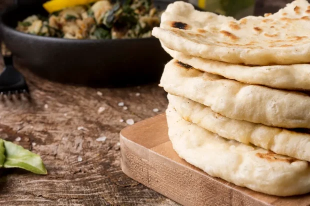 Pilha de pães sírios caseiros, levemente tostados, dispostos sobre uma tábua de madeira rústica, com um prato de acompanhamento ao fundo.