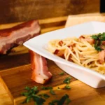 Prato de espaguete à carbonara com pedaços de bacon crocante, decorado com salsa fresca, servido ao lado de uma tábua com bacon defumado e queijo parmesão.