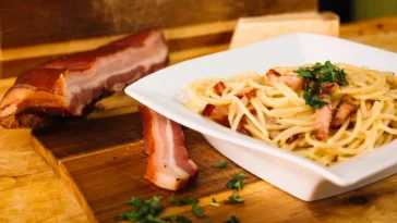 Prato de espaguete à carbonara com pedaços de bacon crocante, decorado com salsa fresca, servido ao lado de uma tábua com bacon defumado e queijo parmesão.