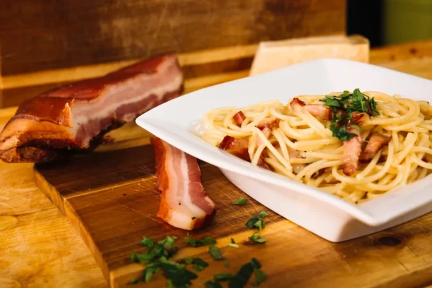 Prato de espaguete à carbonara com pedaços de bacon crocante, decorado com salsa fresca, servido ao lado de uma tábua com bacon defumado e queijo parmesão.