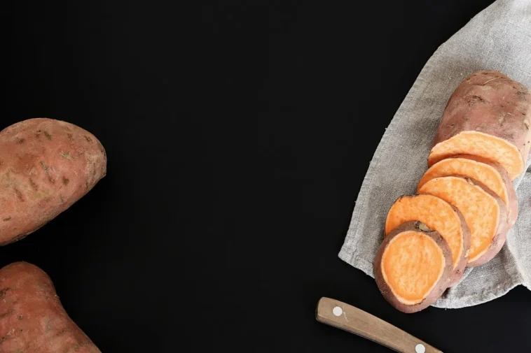 Batata-doce inteira e fatiada sobre um pano de linho cinza com fundo preto minimalista, acompanhada de uma faca de cabo de madeira.