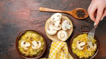 Pratos de cerâmica com cogumelos gratinados, decorados com fatias de cogumelo fresco, ao lado de uma colher de madeira com pimenta e uma toalha amarela.