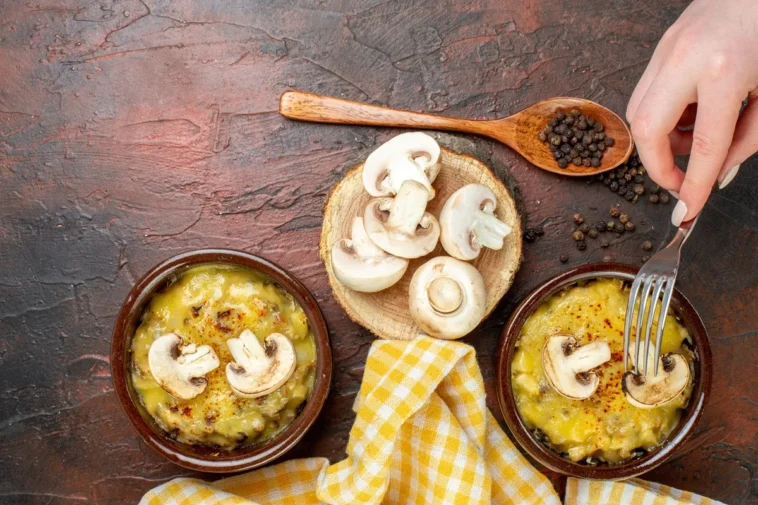 Pratos de cerâmica com cogumelos gratinados, decorados com fatias de cogumelo fresco, ao lado de uma colher de madeira com pimenta e uma toalha amarela.