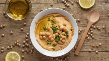 Hummus caseiro servido em tigela branca, decorado com grão-de-bico, salsinha e azeite, acompanhado de limões fatiados, uma colher de madeira e grãos de grão-de-bico espalhados sobre mesa de madeira rústica.