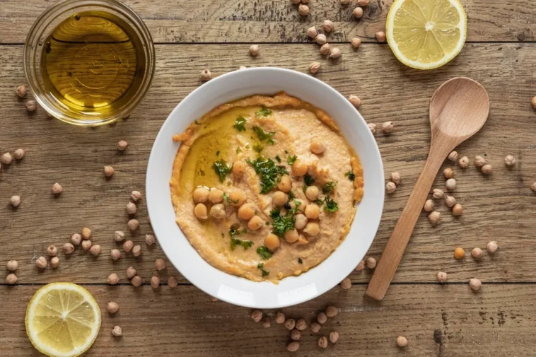 Hummus caseiro servido em tigela branca, decorado com grão-de-bico, salsinha e azeite, acompanhado de limões fatiados, uma colher de madeira e grãos de grão-de-bico espalhados sobre mesa de madeira rústica.