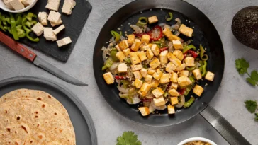 Imagem de uma refeição saudável com cubos de tofu temperado, pimentões verdes, cebolas e quinoa em uma frigideira, acompanhada por tortilhas e ingredientes frescos como coentro e abacate.