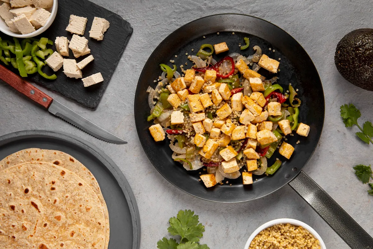 Imagem de uma refeição saudável com cubos de tofu temperado, pimentões verdes, cebolas e quinoa em uma frigideira, acompanhada por tortilhas e ingredientes frescos como coentro e abacate.