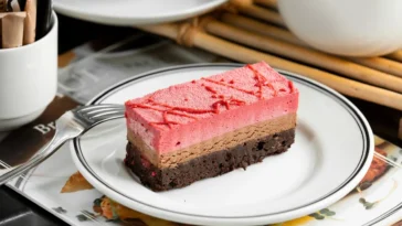 Fatia de bolo gelado com camadas de chocolate, mousse de frutas vermelhas e base de brownie, servida em um prato branco com detalhes pretos.