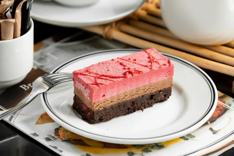 Fatia de bolo gelado com camadas de chocolate, mousse de frutas vermelhas e base de brownie, servida em um prato branco com detalhes pretos.